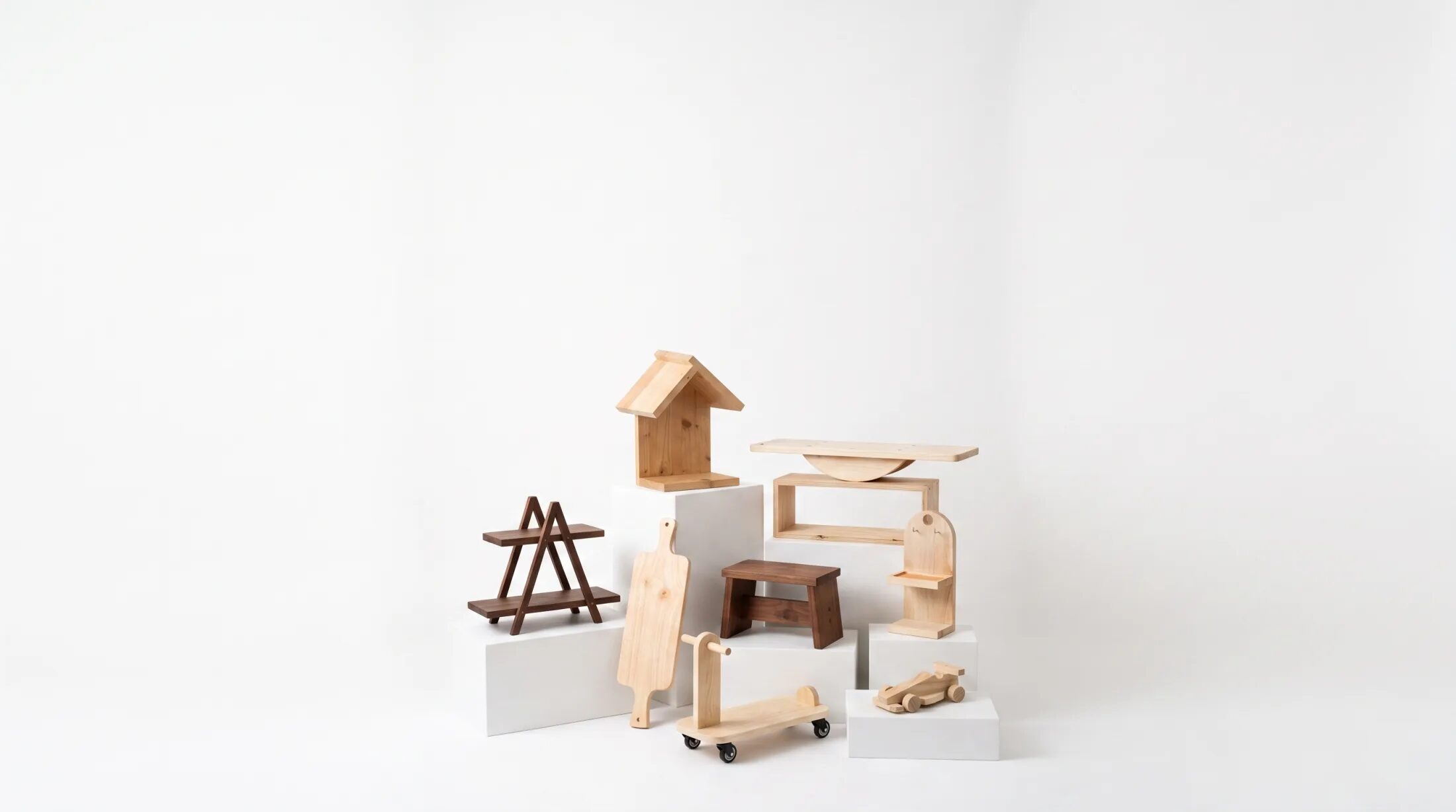 Nine finished Pesta Bina projects grouped on white plinths against a white studio backdrop: a birdhouse, an A-frame shelf, a cutting board, a small seat, a balance board, a scooter, a toy car and a small arched shelf